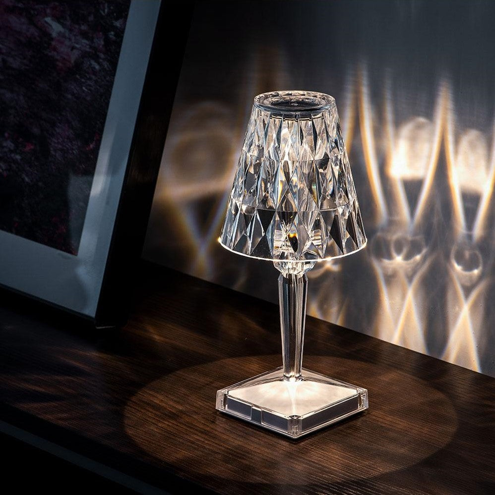 Celestial Glow crystal table lamp with silver finish, illuminated on a dark wooden surface, casting intricate light patterns on wall surface, next to a framed picture, highlighting its decorative effect.