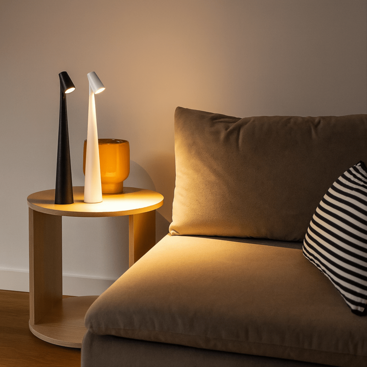 A black and a white Luxe Glow rechargeable table lamp, both illuminated, placed on a round wooden side table next to a cozy sofa, highlighting their modern design and ambient lighting.