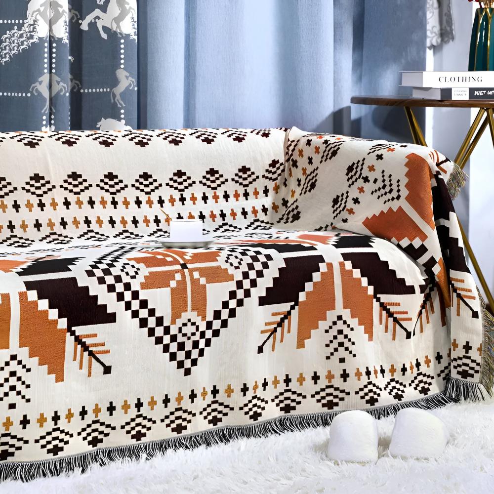 Nomad Oasis picnic blanket with Moroccan boho pattern draped over a sofa, with white slippers on a fluffy rug and a side table with books, creating a cozy indoor setting.