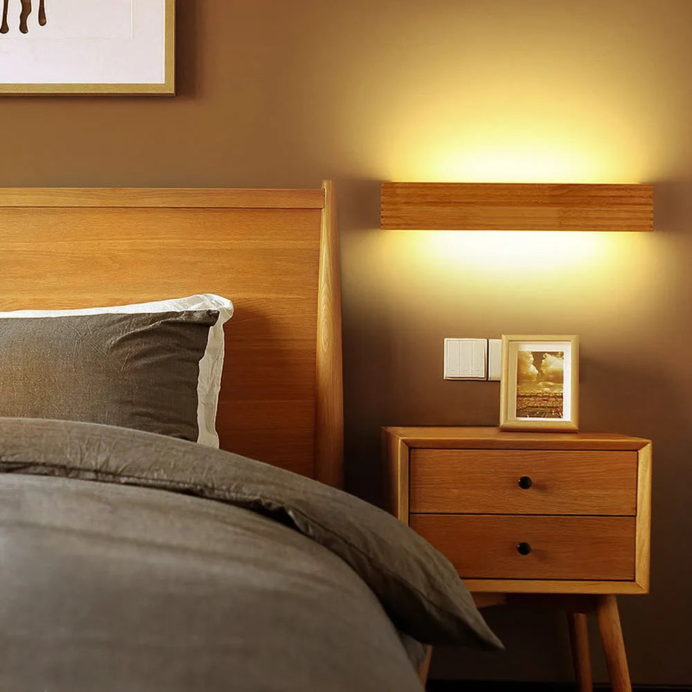 Arctic Glow wood LED wall light mounted above a wooden nightstand in a modern minimalist bedroom, casting a warm glow over the grey bedding.