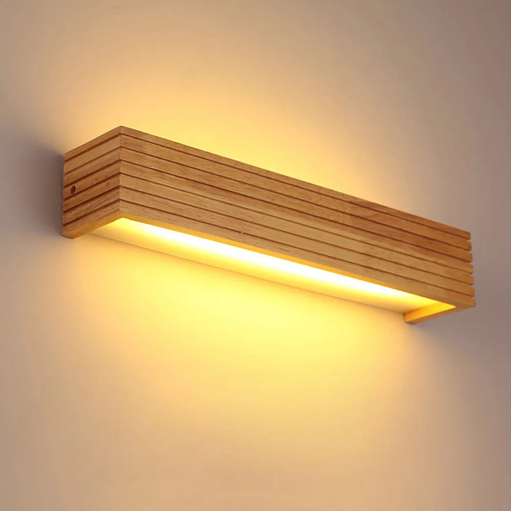 A rectangular wooden LED wall light with a modern minimalist design, mounted on a wall and emitting a warm, soft glow. The light fixture features horizontal wooden slats, creating an inviting ambiance.