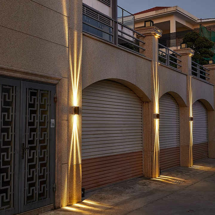 Low angle view of black aluminum three-beam LED wall sconce lit with warm light along outdoor walkway showing black finish