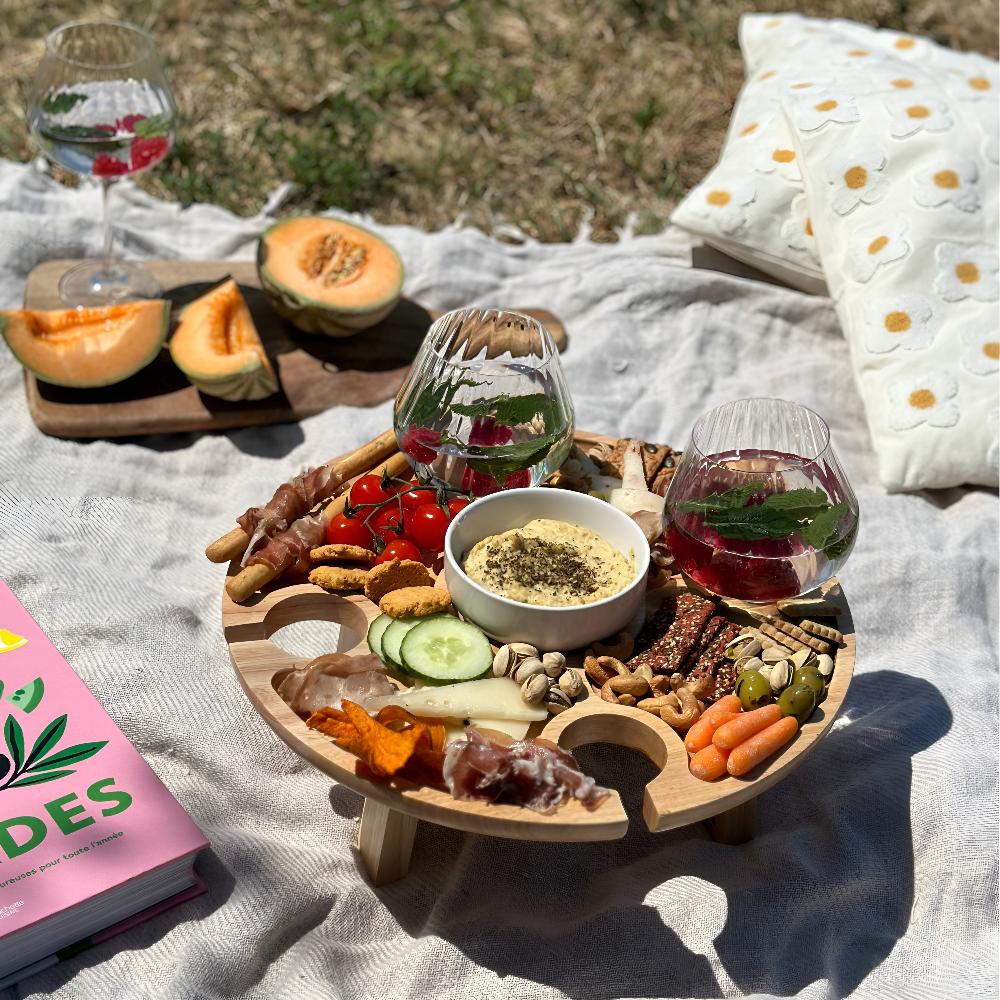 Elysian Gather foldaway picnic table set on a blanket outdoors on grass, featuring a spread of hummus, vegetables, crackers, prosciutto, melon, and nuts, with two glasses of drinks and a book nearby.