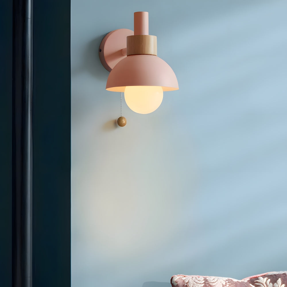 Elysian Glow Classic Wall Sconce in pink with a brushed metal fixture, wooden accent, spherical glass shade, and pull chain switch, emitting warm light in a bedroom setting.