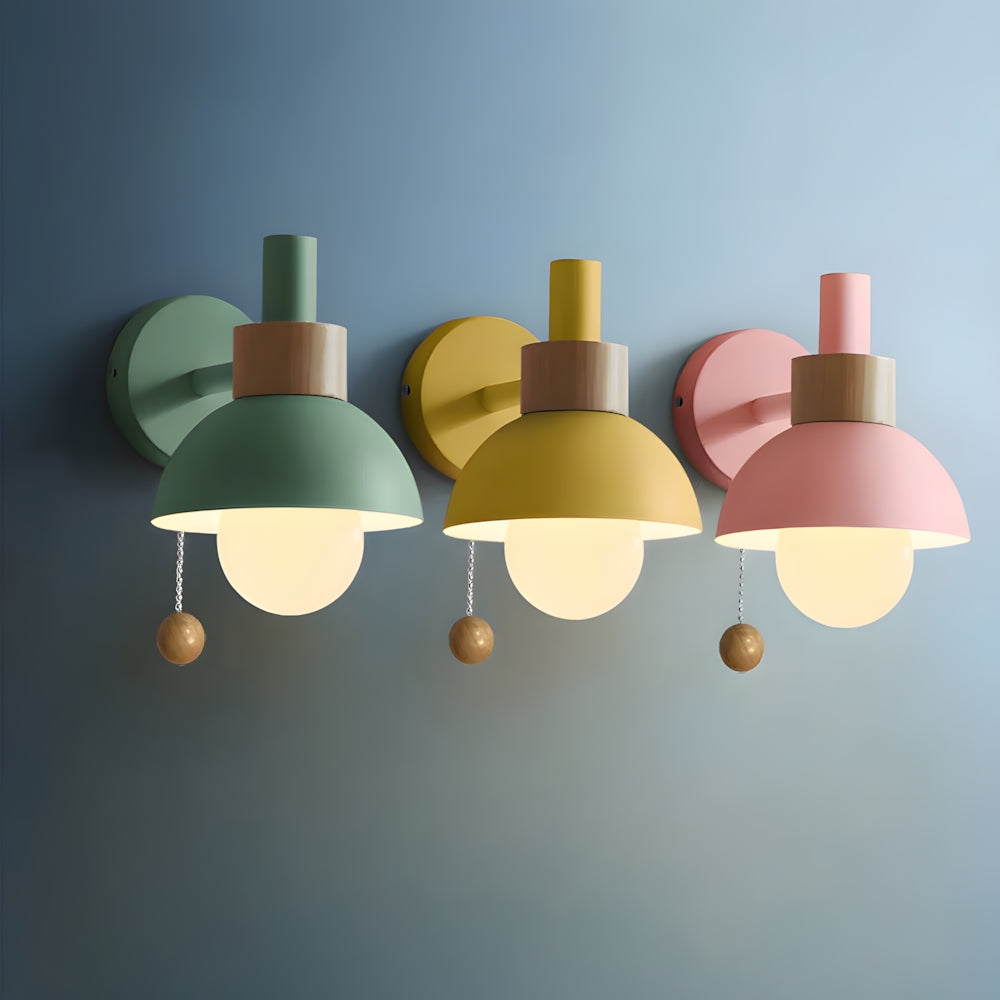 Three Elysian Glow Classic Wall Sconces in green, lemon yellow, and pink, featuring brushed metal fixtures, wooden accents, spherical glass shades, and pull chain switches, emitting warm light on a blue wall.