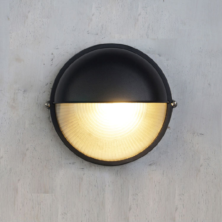 Fenwick vintage outdoor glass sconce wall lamp, black round design, half-illuminated with ribbed glass, mounted on a light wall.