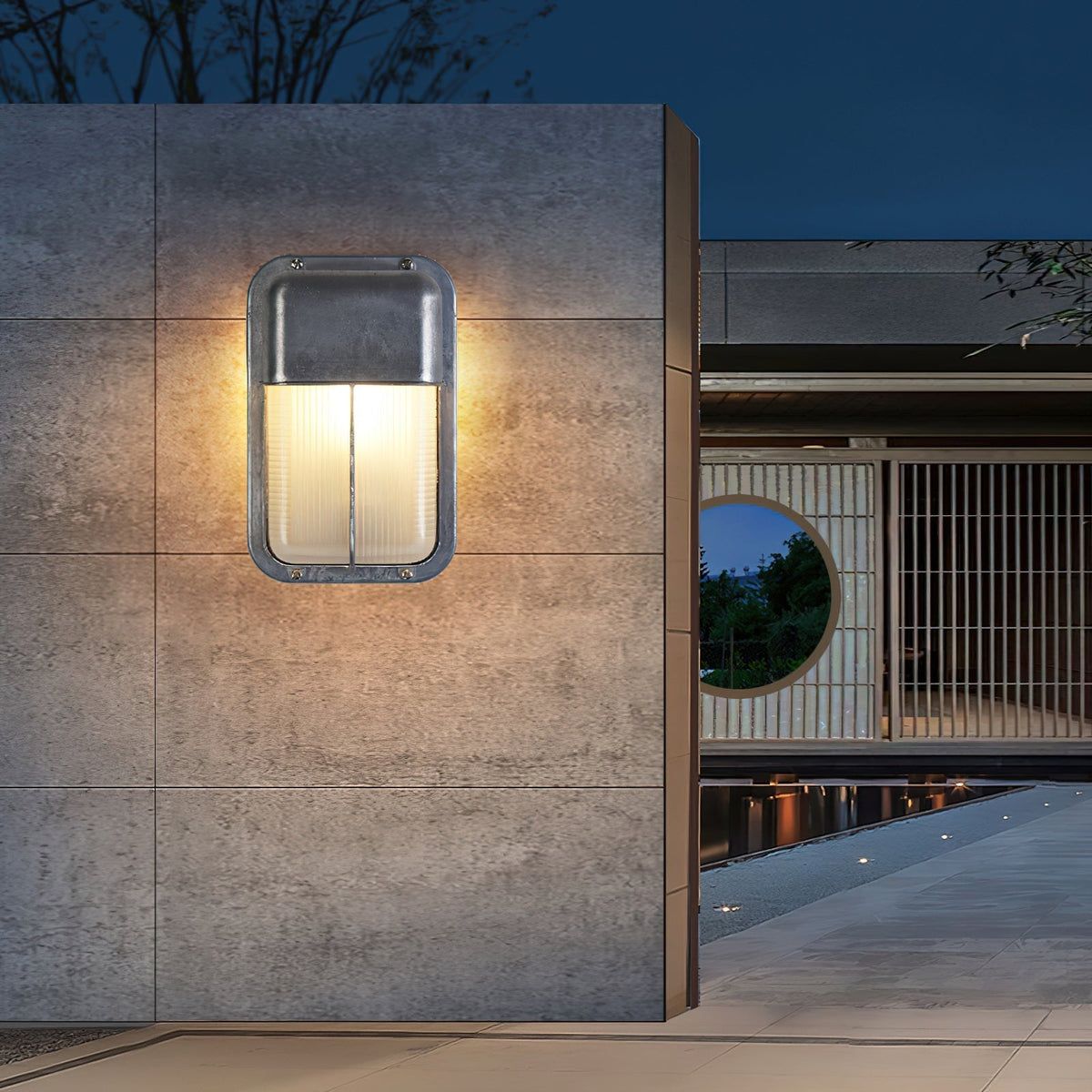 Vintage outdoor glass sconce wall lamp with metal frame and clear glass, illuminated at dusk on a modern grey tiled wall next to a water feature.