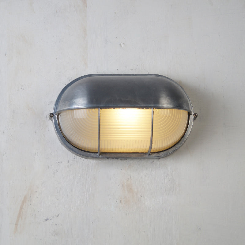 Fenwick vintage outdoor glass sconce wall lamp, featuring an oval-shaped metal frame and clear ribbed glass, illuminated and mounted on a textured light wall.