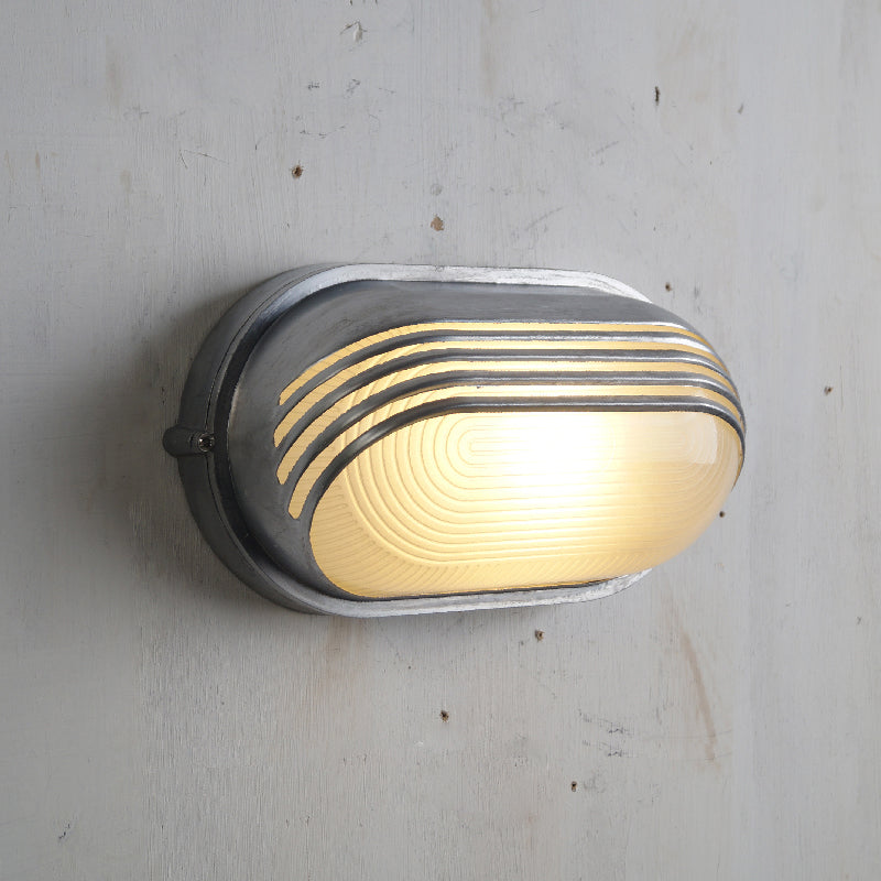 Fenwick vintage outdoor glass sconce wall lamp, oval industrial style metal frame with clear ribbed glass diffuser and horizontal bars, illuminated on a light wall.