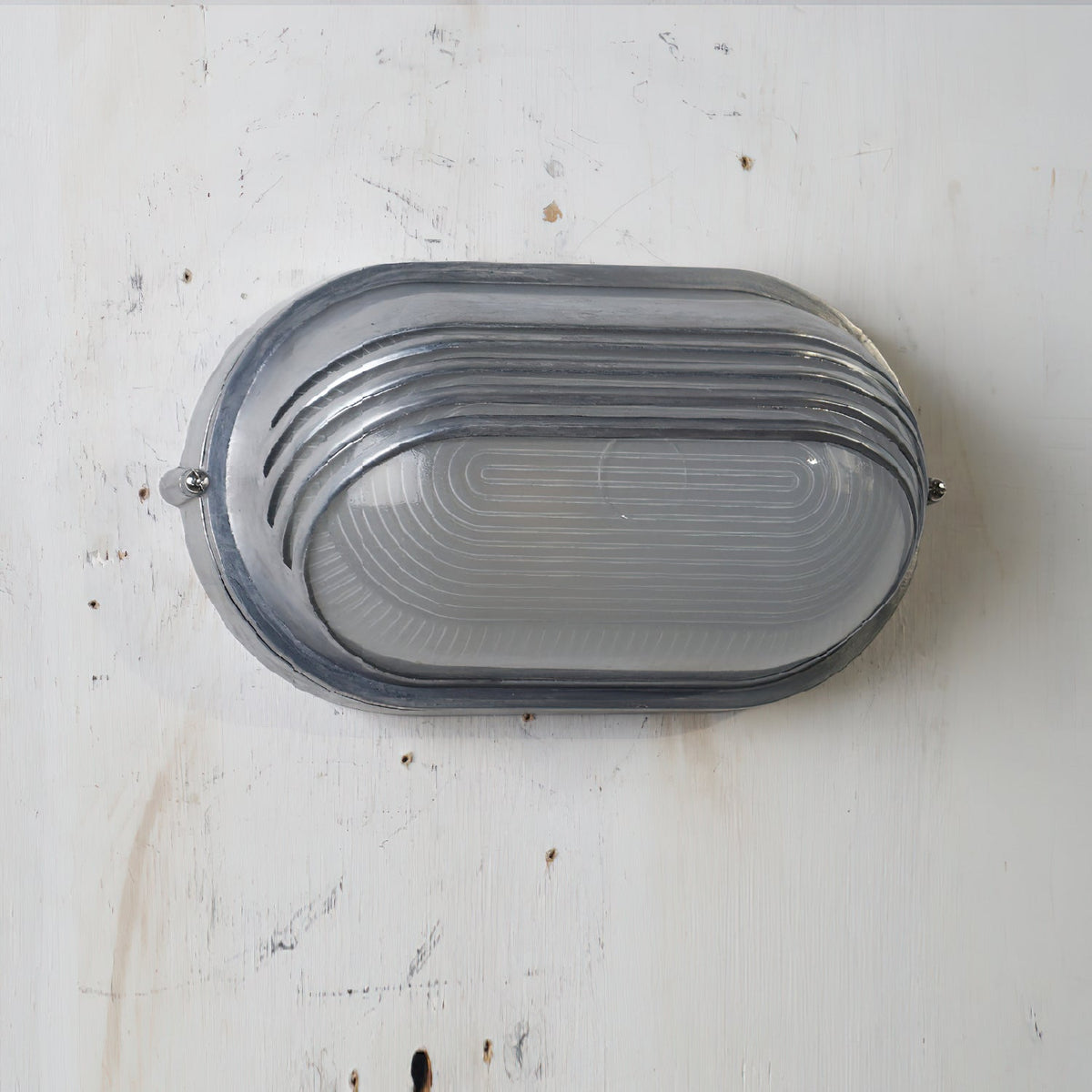 Vintage outdoor glass sconce wall lamp, oval silver metal frame with horizontal ridges and clear glass, mounted on a white wall.