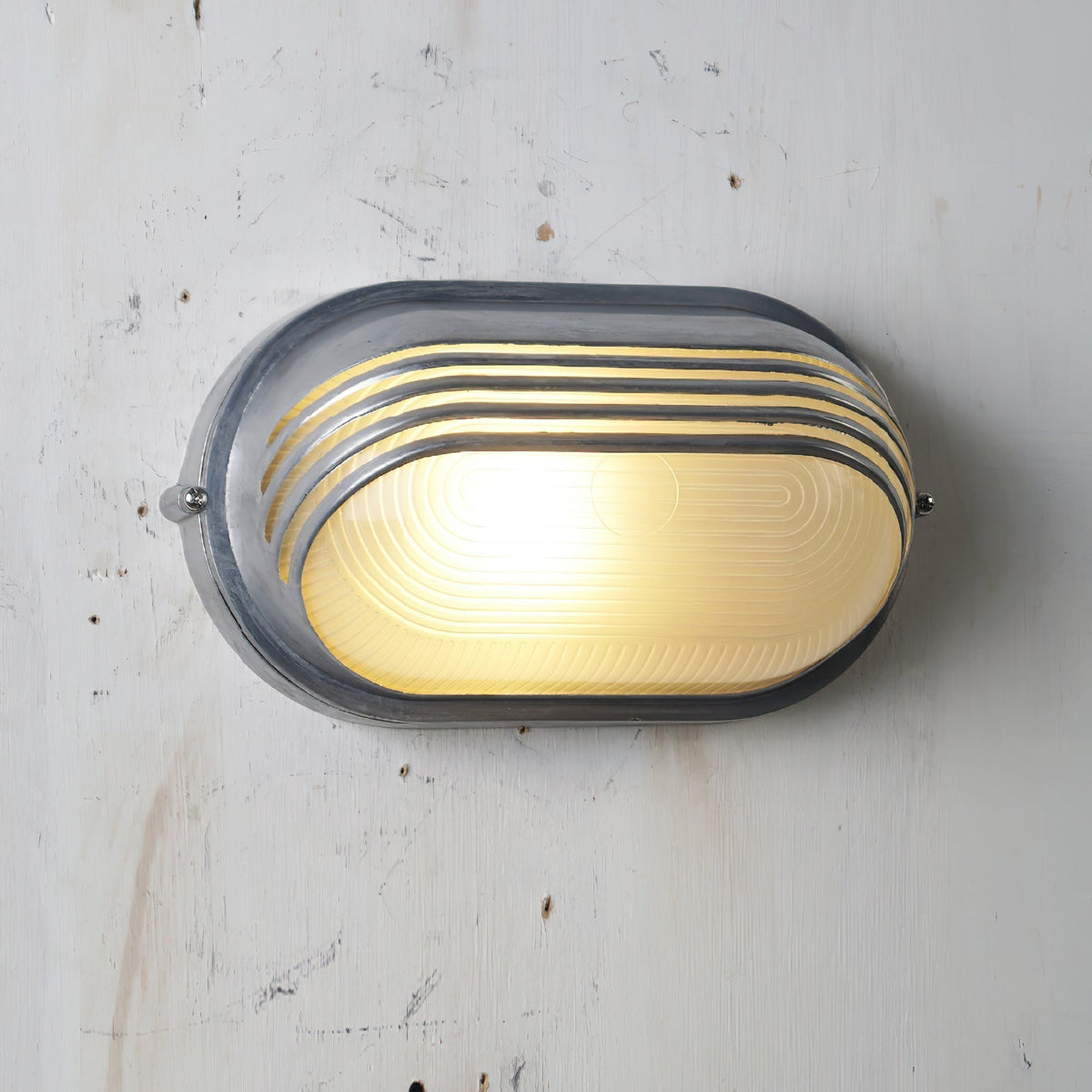 Vintage outdoor glass sconce wall lamp, oval silver metal frame with horizontal ridges and clear glass, illuminated with warm yellow light, mounted on a white wall.