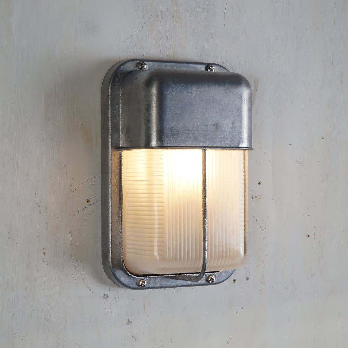 Vintage outdoor glass sconce wall lamp with a rectangular brushed metal frame and ribbed clear glass, illuminated and mounted on a white wall.