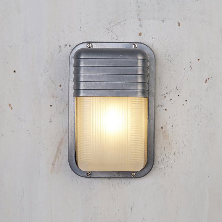 Vintage outdoor wall sconce with a rectangular, dark metal frame featuring horizontal ridges and ribbed clear glass diffuser, illuminated on a light grey textured wall.