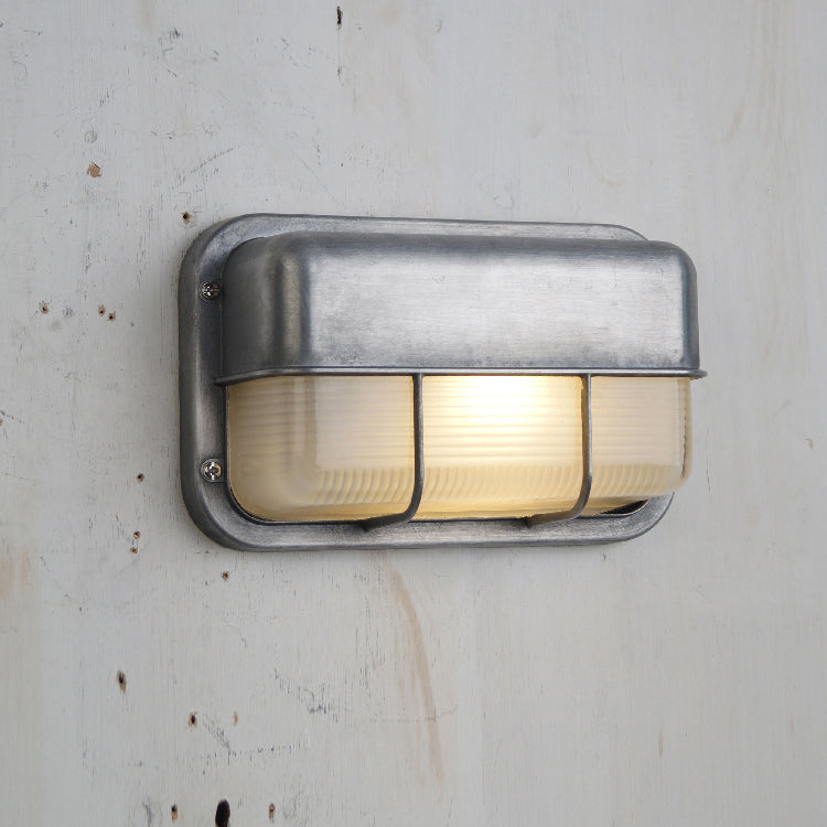 Close-up of a Fenwick vintage outdoor glass sconce wall lamp, featuring a rectangular metal frame with a brushed finish and clear ribbed glass, illuminated and mounted on a textured light wall.