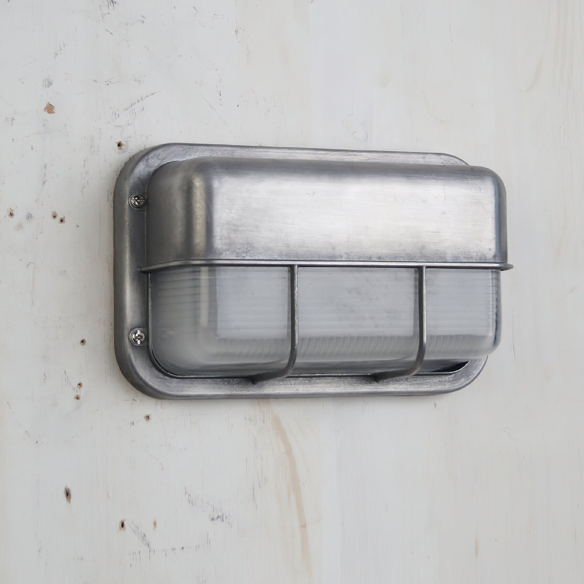 Vintage outdoor wall sconce with a rectangular metal frame, clear ribbed glass, and a protective metal cage, shown unlit on a light wooden background.