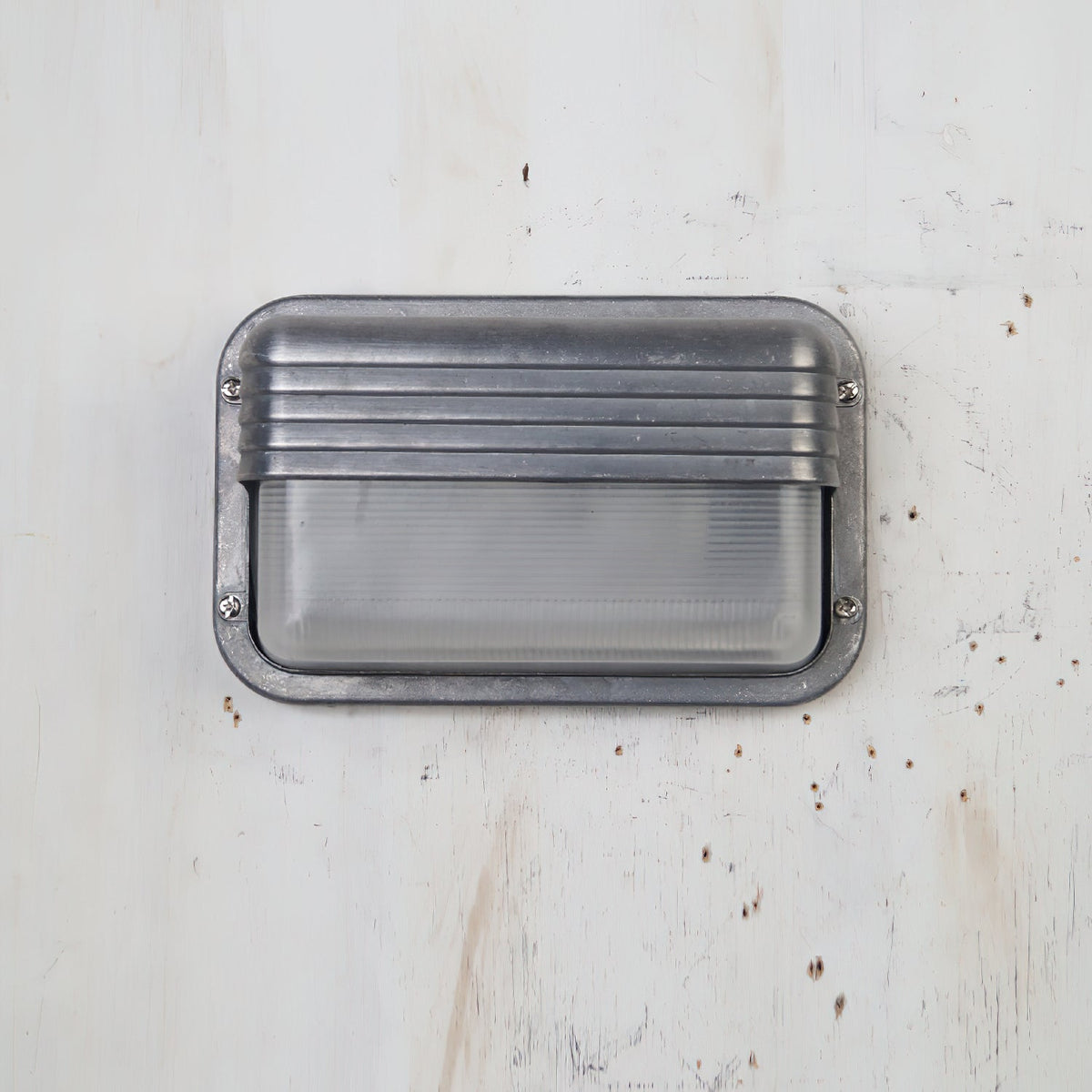 Vintage outdoor wall sconce with a rectangular metal frame featuring horizontal ridges, clear ribbed glass, shown unlit without a cage on a light wooden background.