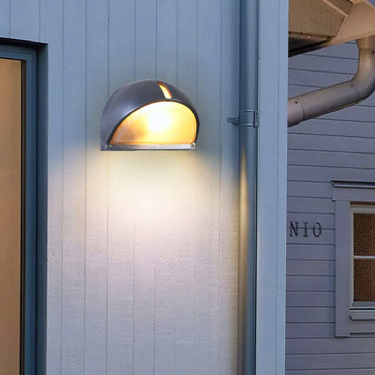 Fenwick vintage outdoor wall sconce, silver half-dome metal frame with clear glass, illuminated and mounted on a white exterior wall next to a window.