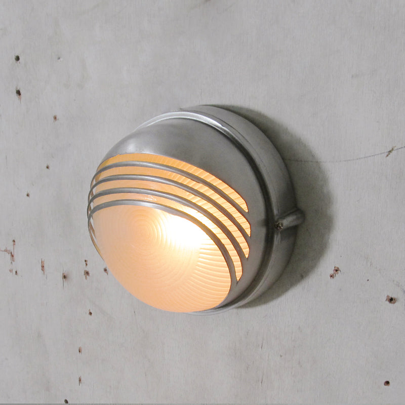 Fenwick vintage outdoor wall sconce, silver round metal frame with horizontal bars and clear ribbed glass diffuser, illuminated on a textured grey wall.