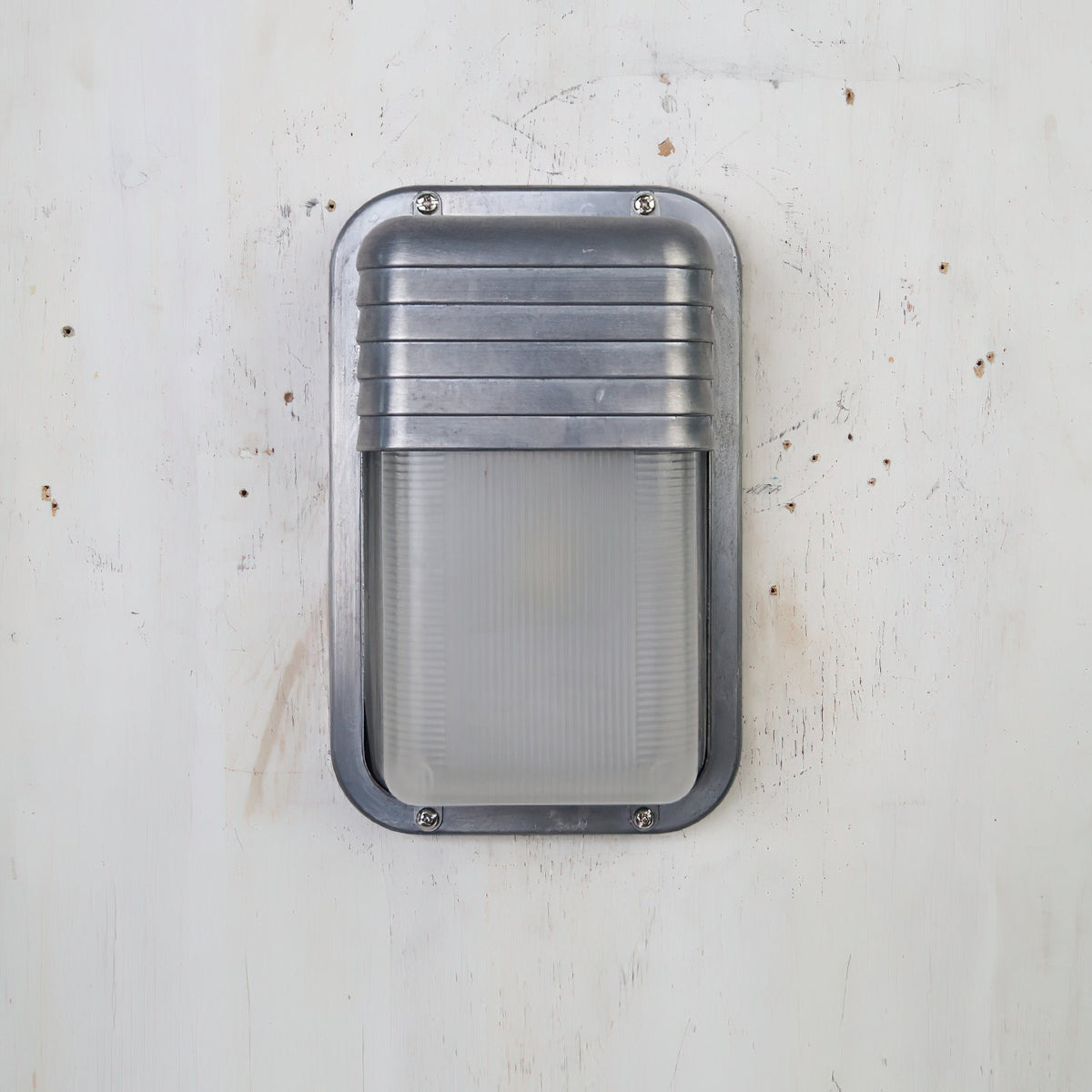 Fenwick vintage outdoor glass sconce wall lamp with a silver metal frame, ribbed metal top, and clear ribbed glass diffuser, shown off on a distressed white wall.