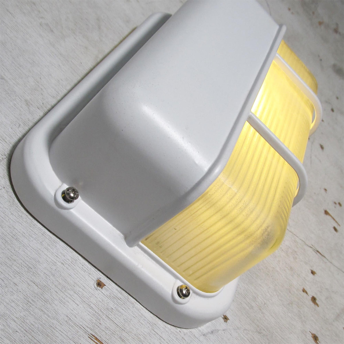 Angled view of Fenwick vintage outdoor glass sconce wall lamp with a white metal frame and clear ribbed glass diffuser, shown on with a warm yellow glow.
