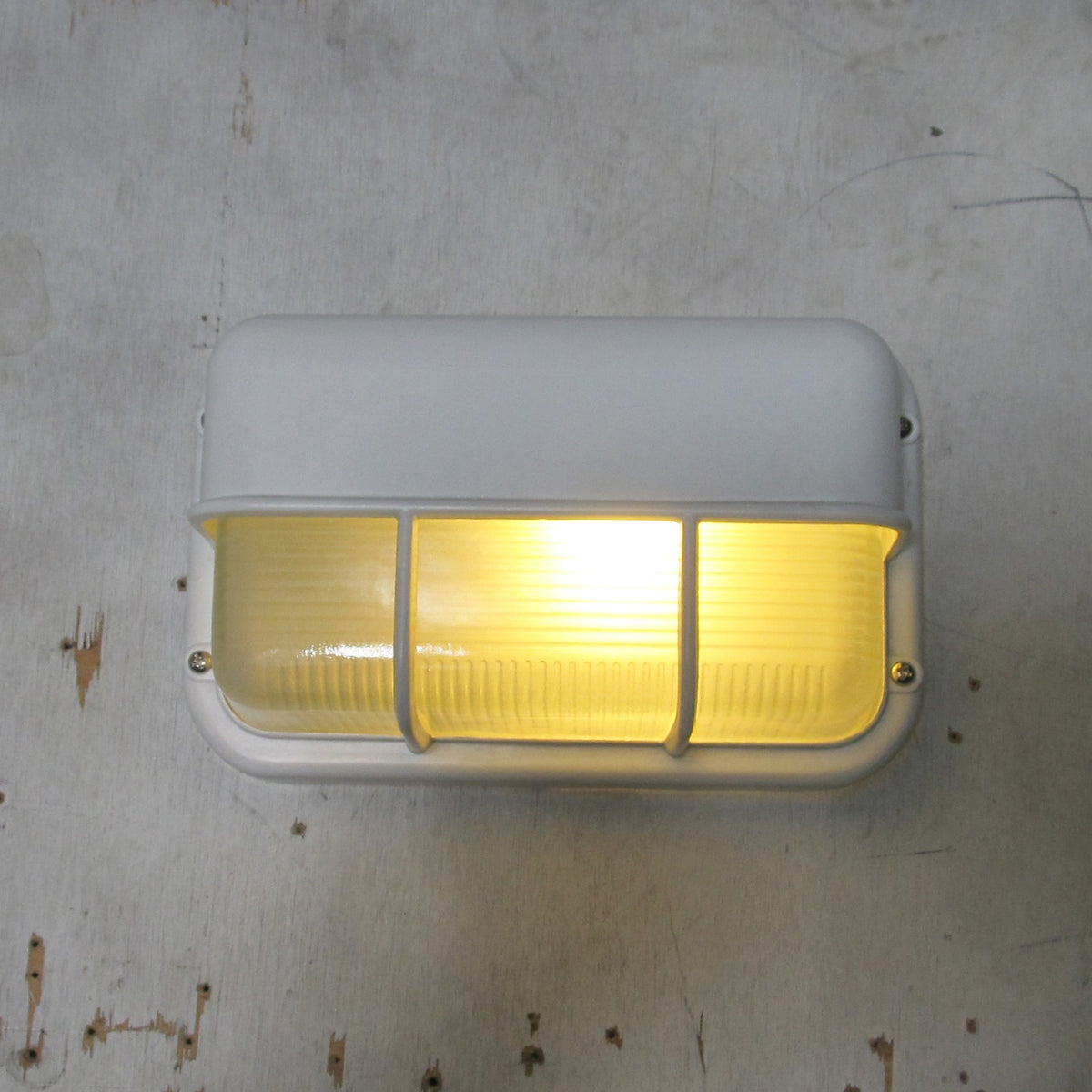 Fenwick vintage outdoor glass sconce wall lamp with a white metal frame and clear ribbed glass diffuser, shown on with a warm yellow glow on a distressed white wall.