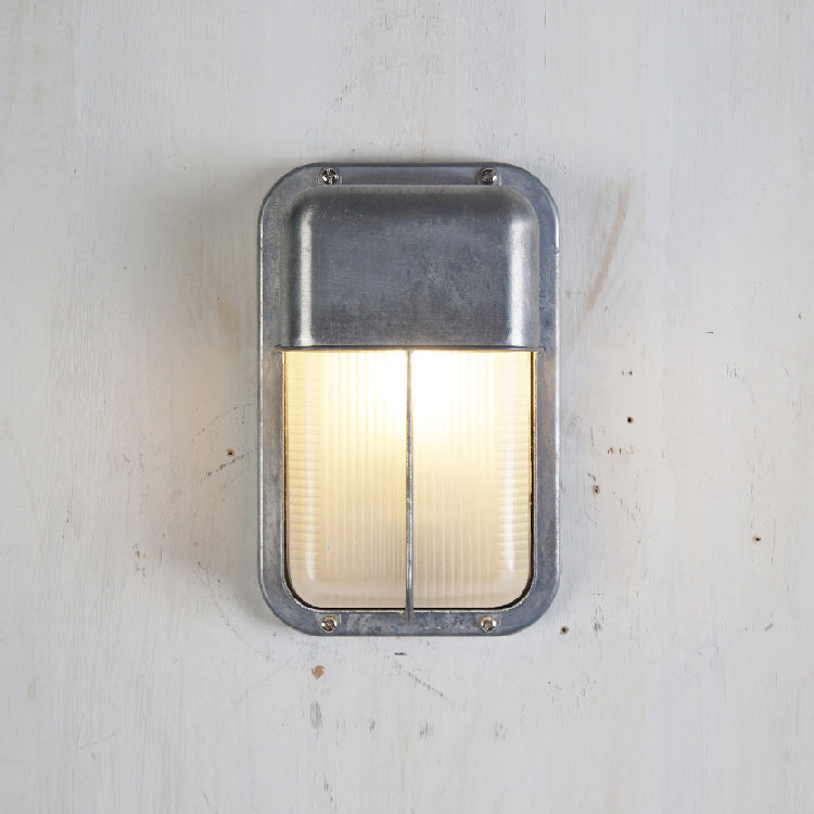 Fenwick vintage outdoor glass sconce wall lamp with rectangular silver metal frame and ribbed clear glass, glowing, mounted on a white textured wall.