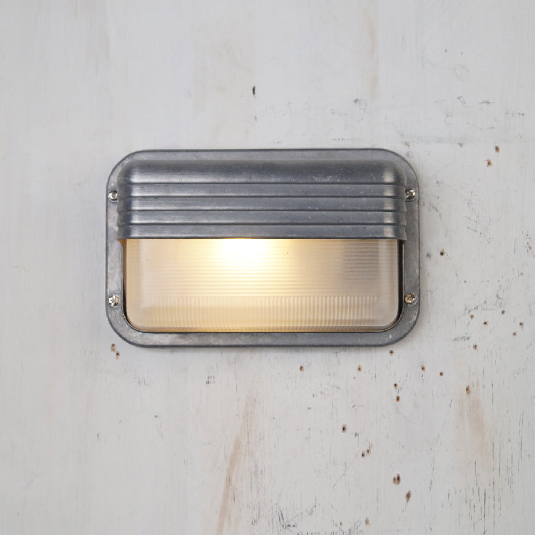Fenwick vintage outdoor glass sconce wall lamp with rectangular silver metal frame, horizontal ridges, and clear glass, glowing, mounted on a white textured wall.