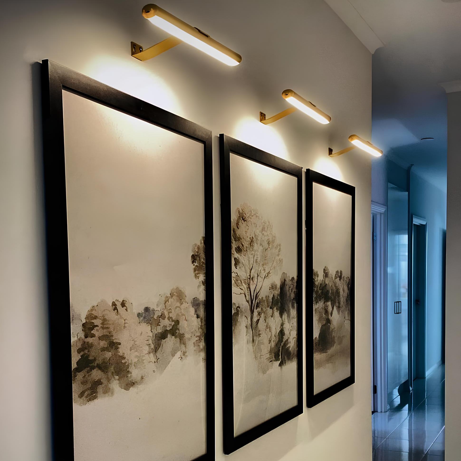 Three gold Luminous Haven Cordless Wall Art Lights with Adjustable Brightness illuminating landscape paintings in a hallway.