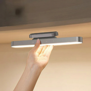 Lumora wireless magnetic LED lamp in gray, held by a hand from underneath, showcasing its illuminated light and control buttons.