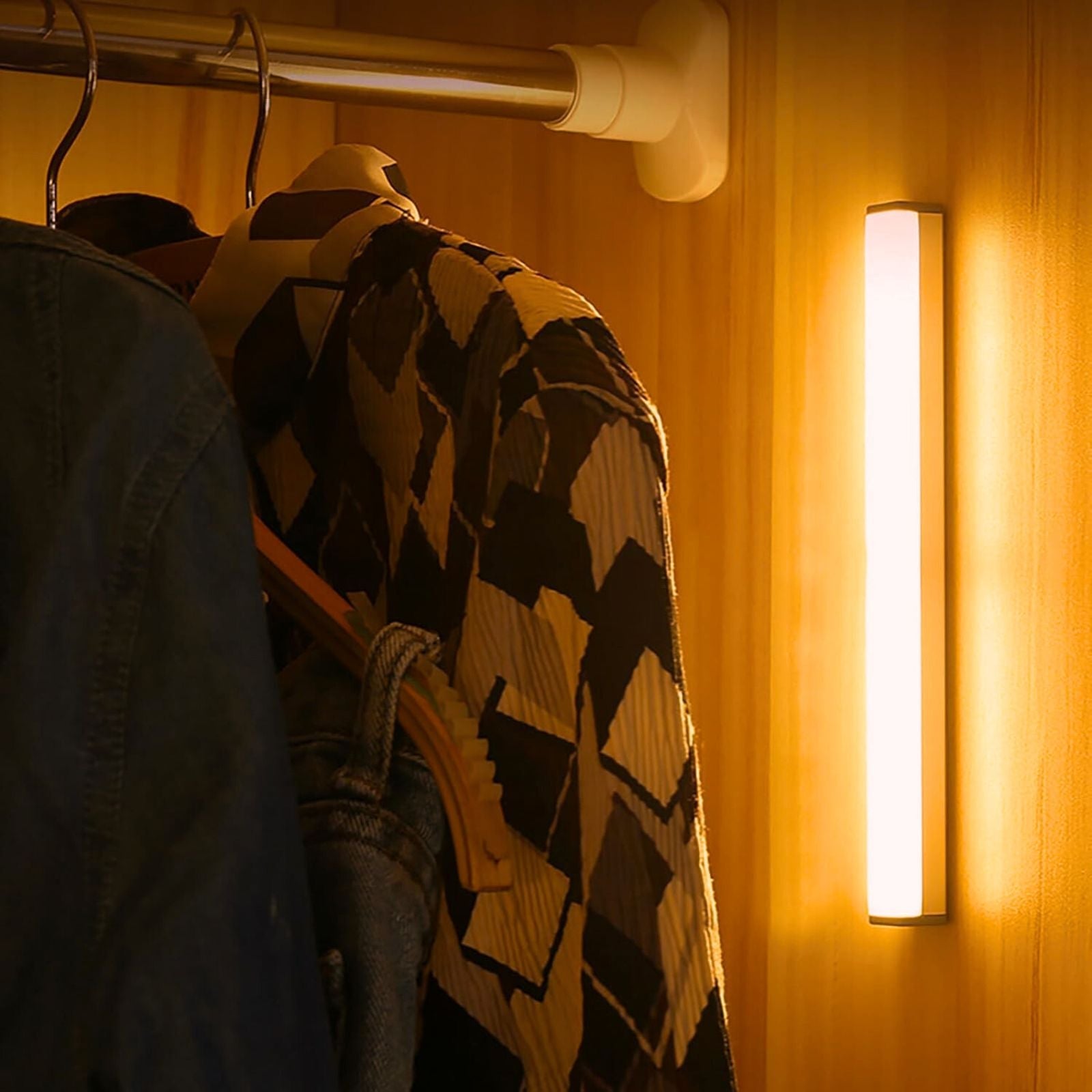 Lumos Aura motion sensor light strip emitting warm white light, mounted vertically inside a wooden closet, illuminating hanging clothes.