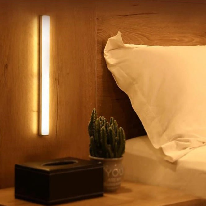 Lumos Aura motion sensor light strip emitting warm white light, mounted vertically on a wooden wall next to a bed, with a cactus plant and tissue box on the bedside table.