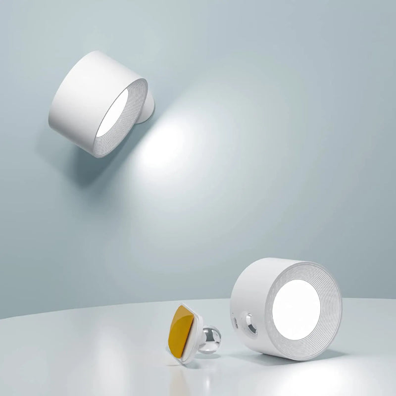 Two white Lumos cordless wall lamps. One lamp is mounted on a light blue wall, illuminating the surface below. The second lamp is shown disassembled on a white table, revealing the magnetic base a yellow accent lamp unit.