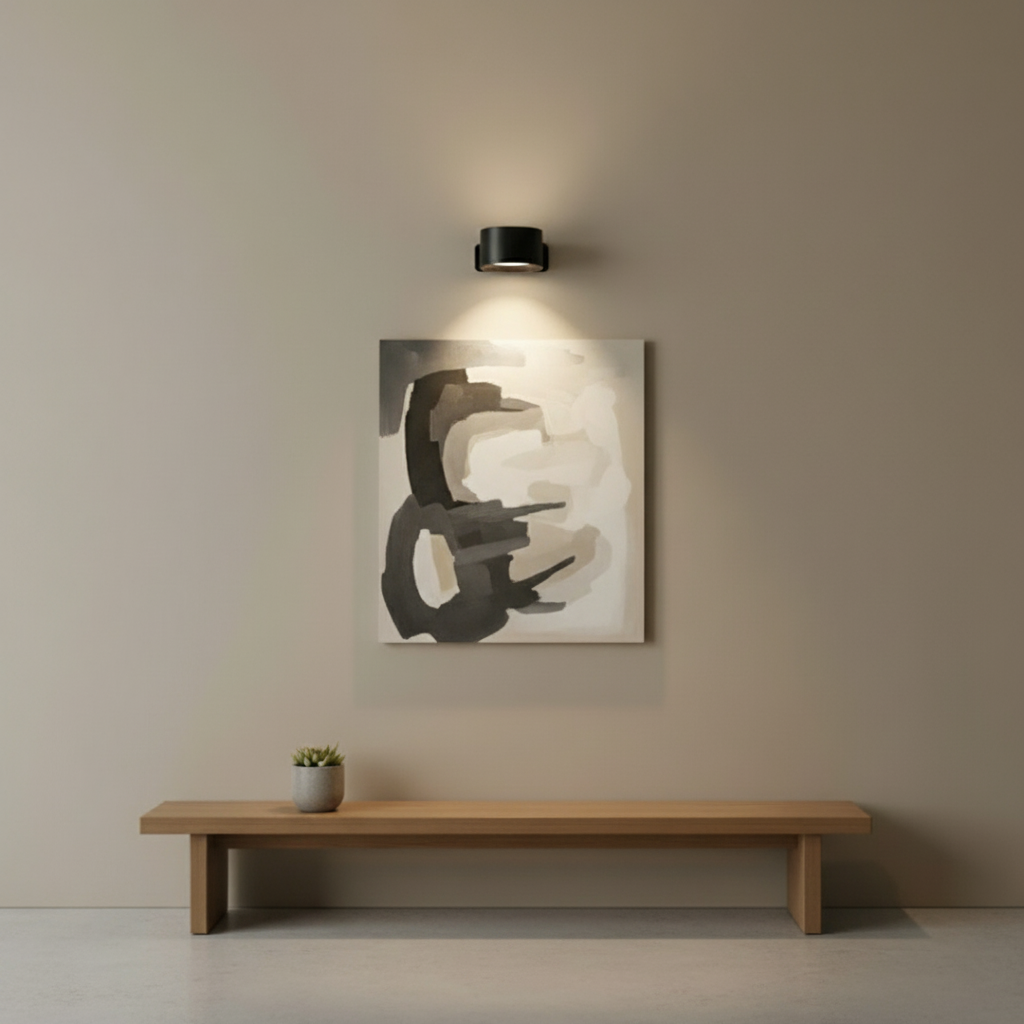 A black Lumos wireless wall lamp with LED lighting mounted on a light beige wall, illuminating an abstract painting and a wooden bench below.