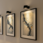 Multiple black Lumos wireless wall lamps mounted above framed abstract artwork on a light-colored wall. Each lamp provides focused LED lighting to highlight details modern art pieces, creating an illuminated art gallery display.