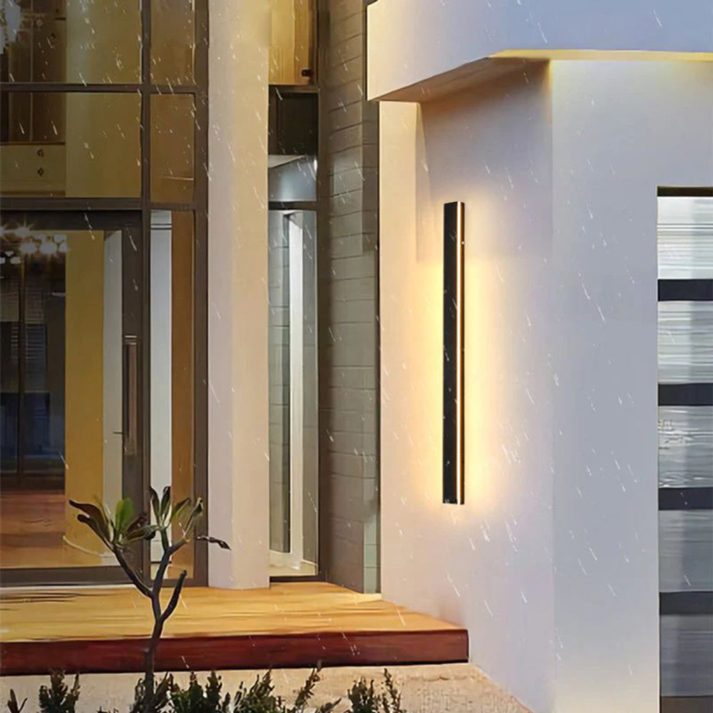 Luna Edge IP65 linear LED wall light in black metal with acrylic finish, emitting warm white light on a modern building exterior during rain.