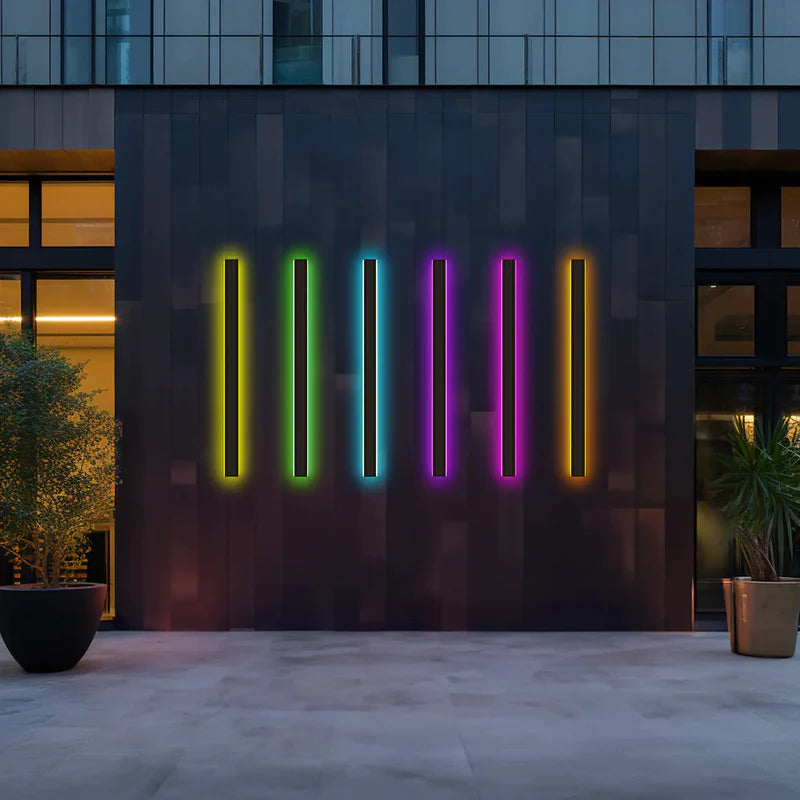 Five Luna Edge IP65 linear LED wall lights displaying vibrant RGB colors (yellow, green, blue, purple, orange) on a dark modern building exterior at dusk.