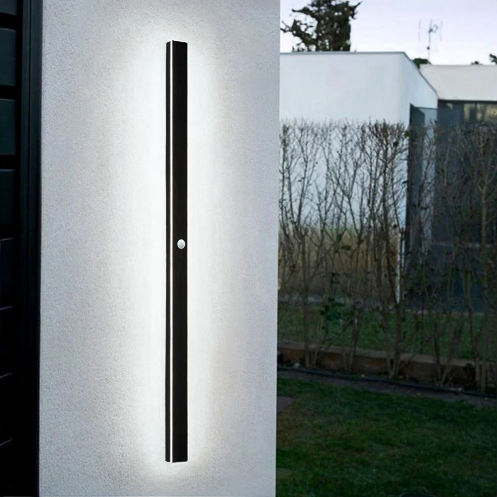Luna Edge IP65 linear LED wall light in black metal with acrylic finish, featuring a motion sensor, installed vertically on a white exterior wall. The sleek outdoor fixture provides bright white illumination, ideal for modern homes and security.