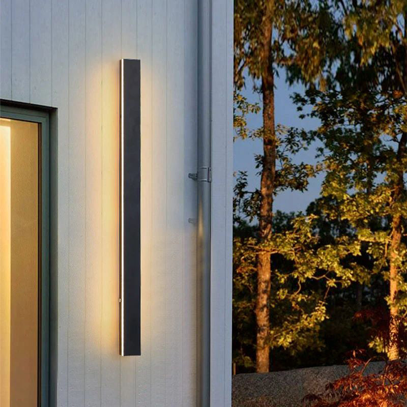 Luna Edge IP65 linear LED wall light in black metal with acrylic finish, installed vertically on a modern white exterior wall, providing warm illumination at dusk. This weather-resistant outdoor light enhances the building's facade and surrounding landscape.