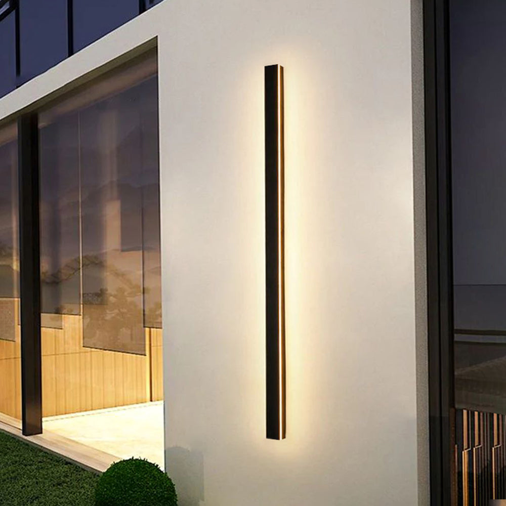 Luna Edge IP65 linear LED wall light in black metal with acrylic finish, casting warm light on a modern white exterior wall, showcasing its sleek design and outdoor illumination.
