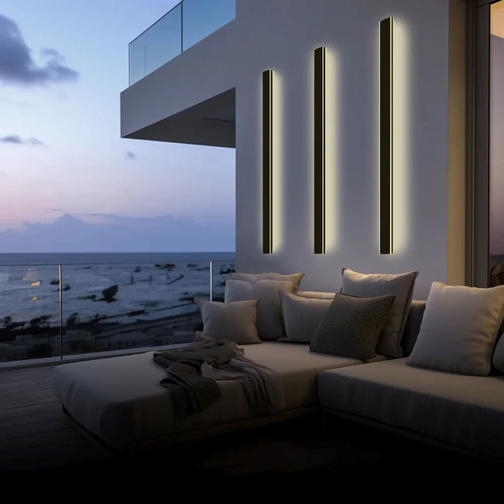 Three Luna Edge IP65 linear LED wall lights in black metal with acrylic finish, providing warm illumination on a modern patio overlooking the ocean at dusk, enhancing the outdoor living space.