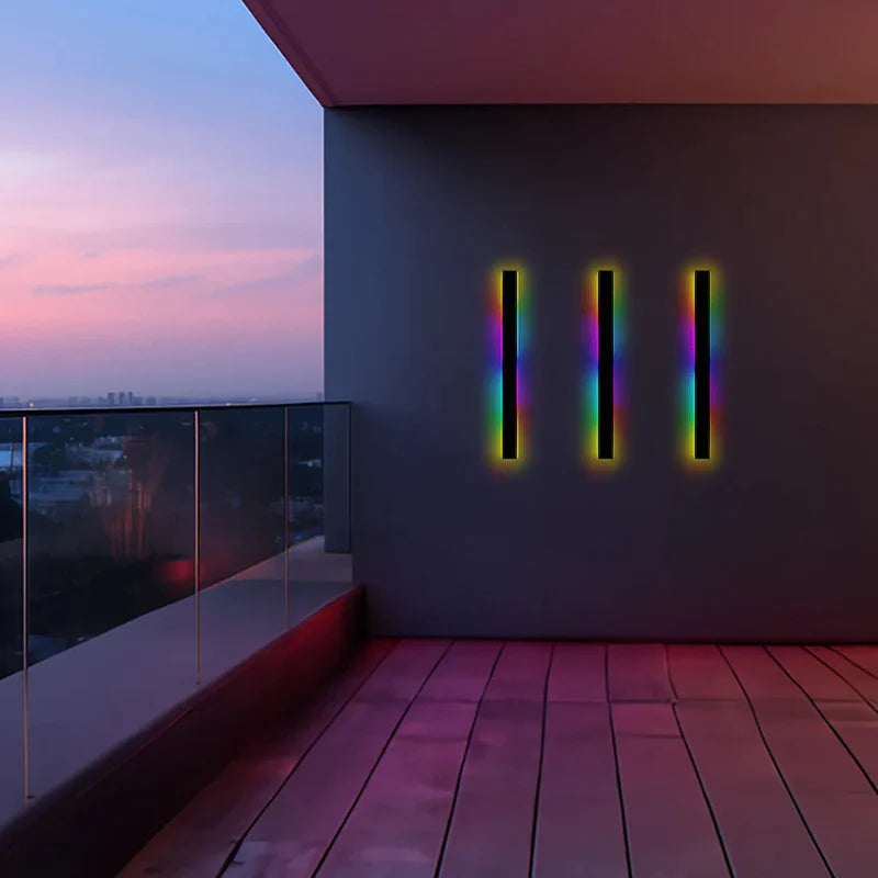 Luna Edge IP65 Linear LED Wall Light in black metal with acrylic finish, showing three RGB lights mounted vertically on an outdoor balcony wall at sunset, illuminating the space with vibrant colors. Modern outdoor lighting fixture, weather resistant.