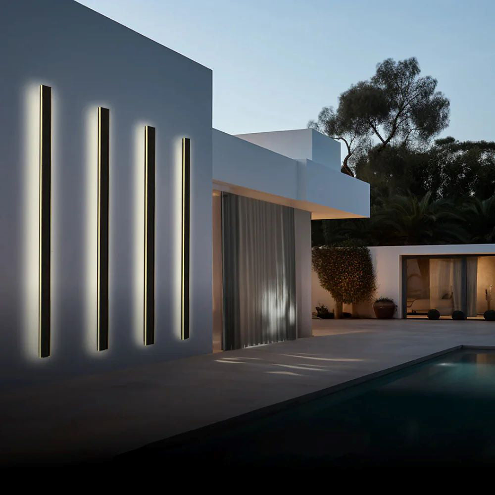 Luna Edge IP65 Linear LED Wall Light in black metal with acrylic finish, showcasing three vertical fixtures emitting warm white light on a modern building exterior next to a swimming pool at dusk. Ideal IP65 rated outdoor wall light.