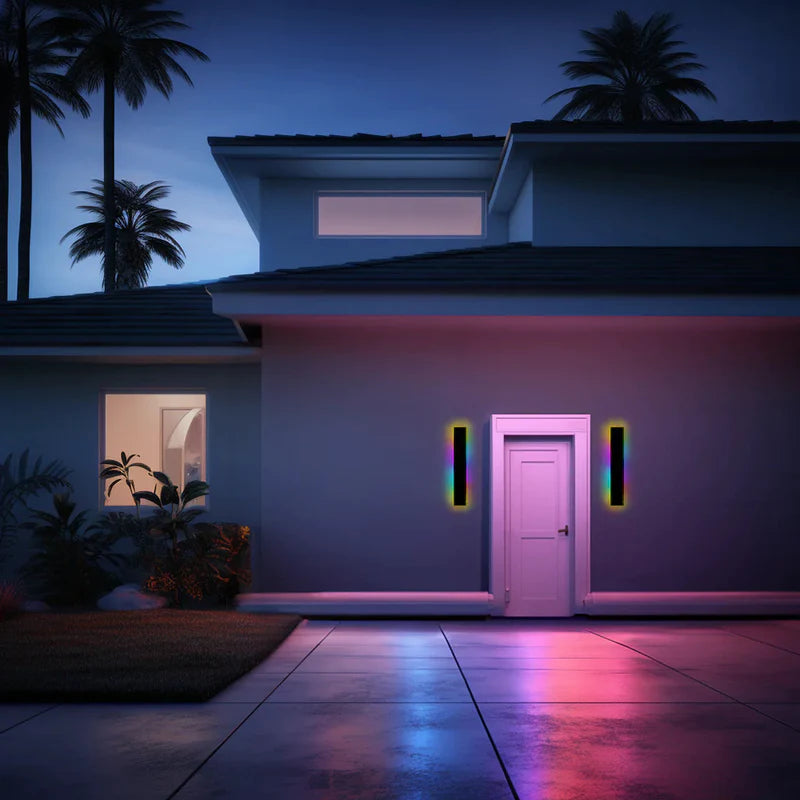 Two Luna Edge IP65 linear LED wall lights in black metal with acrylic finish, displaying RGB colors on either side of a white front door of a modern house at dusk.