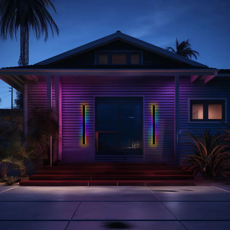 Two Luna Edge IP65 linear LED wall lights in black metal with acrylic finish, displaying RGB colors on either side of a house entrance at dusk.