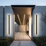 Two Luna Edge IP65 linear LED wall lights in black metal with acrylic finish, installed vertically on either side of a modern building's entrance. These sleek outdoor sconces provide bright, even illumination, enhancing the architectural design and welcoming guests.