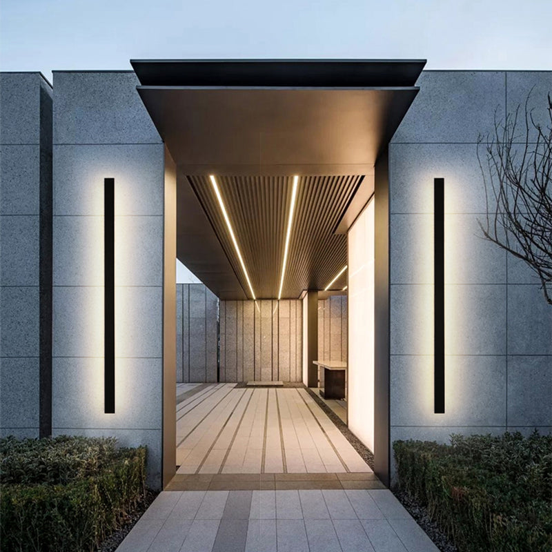 Two Luna Edge IP65 linear LED wall lights in black metal with acrylic finish, installed vertically on either side of a modern building's entrance. These sleek outdoor sconces provide bright, even illumination, enhancing the architectural design and welcoming guests.