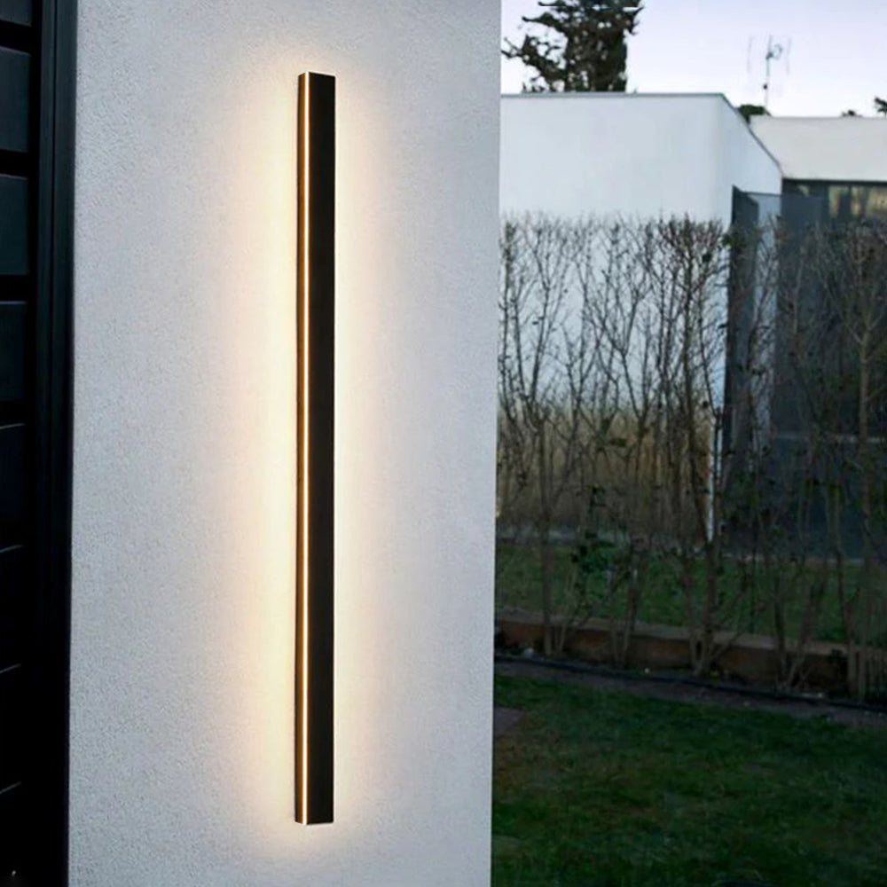 A sleek black metal Luna Edge IP65 linear LED wall light mounted vertically on a modern white exterior wall, casting a warm, inviting glow. In the background, a contemporary house and garden are visible. This weather-resistant fixture provides elegant exterior illumination.