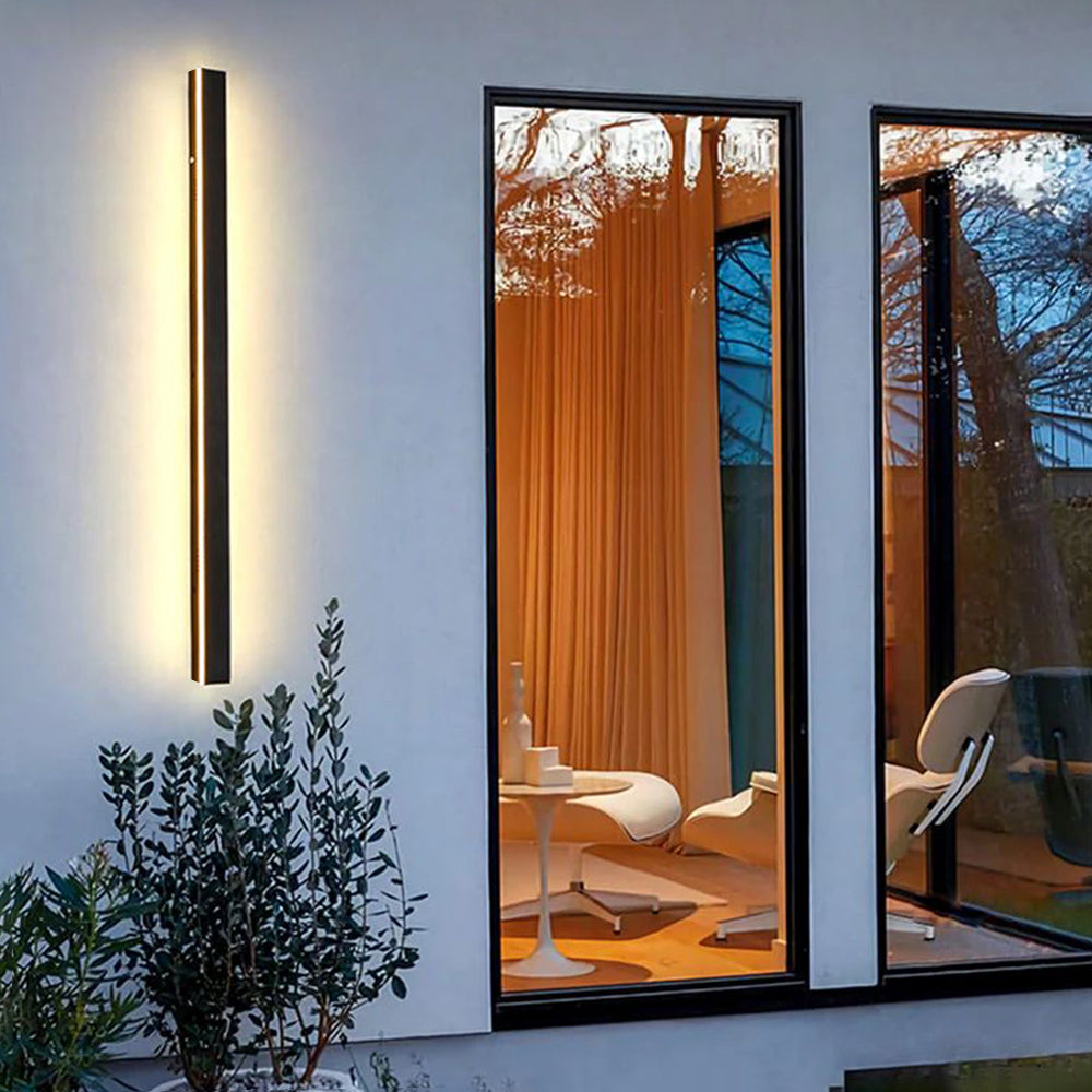 A modern black metal Luna Edge IP65 linear LED wall light mounted vertically on a white exterior wall, illuminating green plants below with a warm glow. Large windows reflect the outdoor environment and show a glimpse of an interior space.