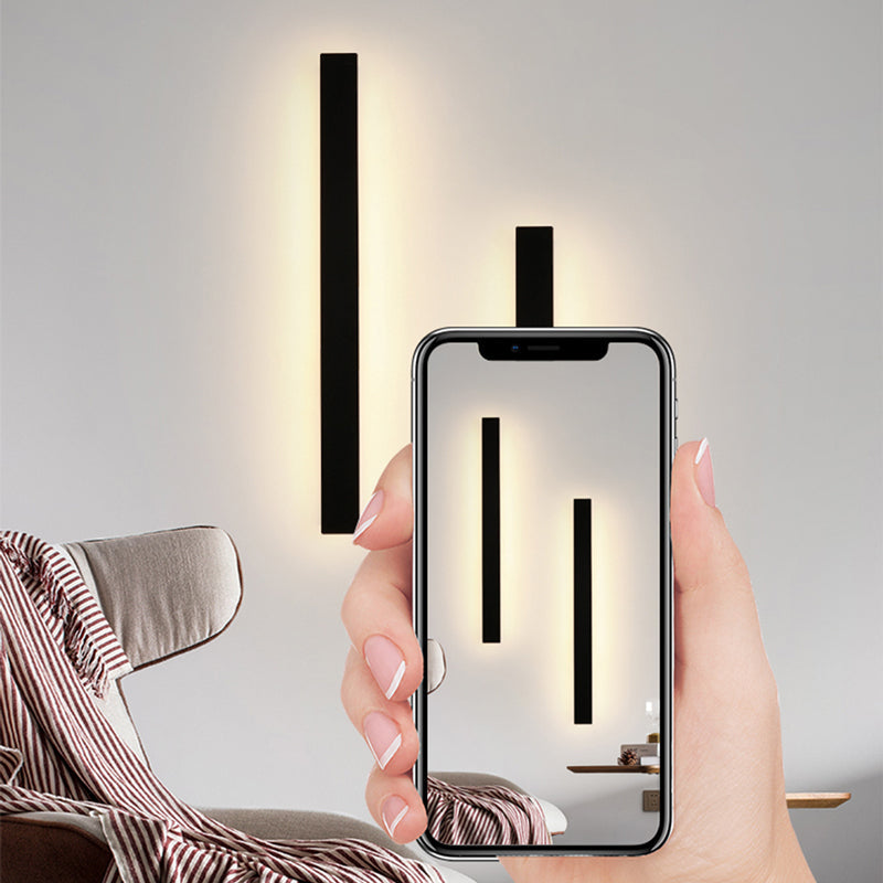 A woman's hand holding a smartphone, reflecting two Luna Edge IP65 linear LED wall lights emitting a warm glow, with one light installed on the wall behind.