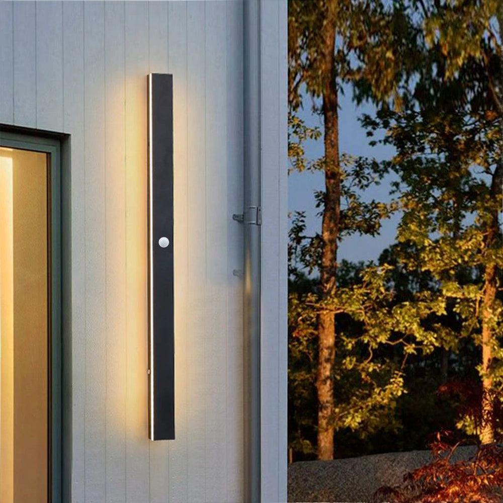 Luna Edge IP65 Linear LED Wall Light Black Metal with Acrylic Finish, showing a tall black linear LED fixture emitting warm white light, mounted vertically on a white exterior wall beside a window. A motion sensor is visible on the light. The image captures the light at dusk with trees in the background, emphasizing its modern outdoor illumination.